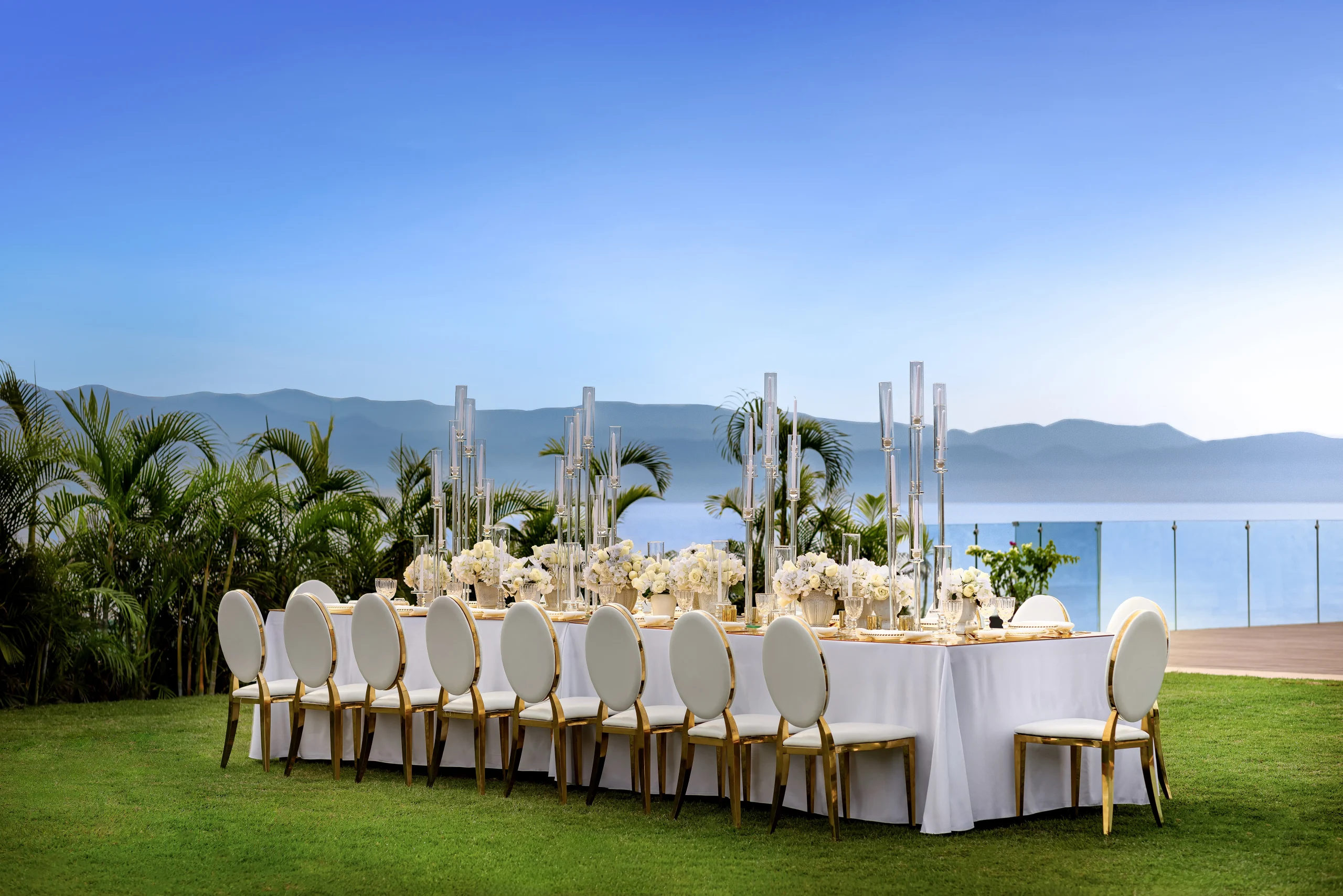 drbmiwedreceptiongardendeck03afcresized680046d430dd2 be all inclusive Outdoor seaside wedding reception with a long white-draped table, white flowers, tall glass candle holders, and palm plants outside.