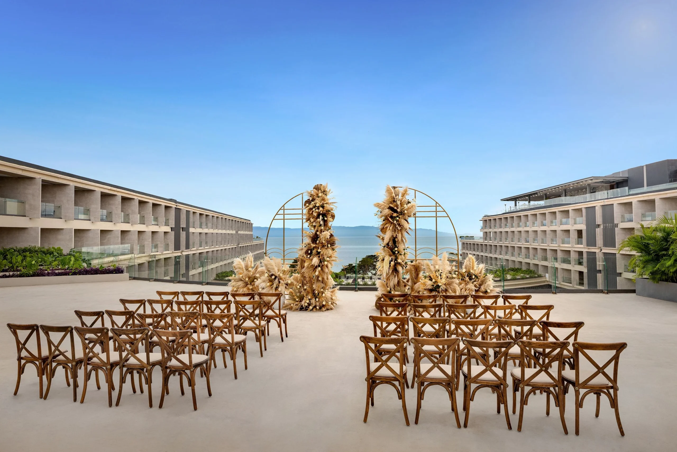 drbmiwedreceptionmoondeckafc6852e1b367df4 be all inclusive Outdoor wedding ceremony set on a terrace, wooden folding chairs arranged in rows facing a pampas grass arch with a sea view.