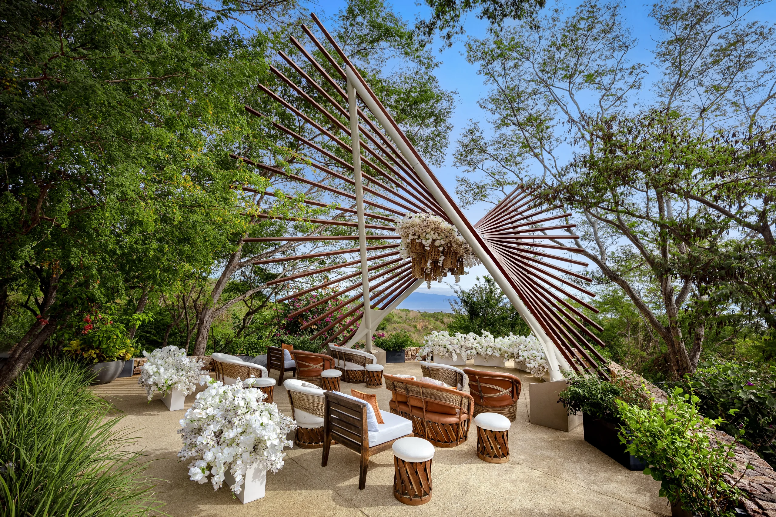 drbmiwedreceptionthenestafcresized680149629737b be all inclusive Outdoor wedding or event seating area with round wicker chairs, white cushions, and tall white floral arrangements under a metal sunburst sculpture.