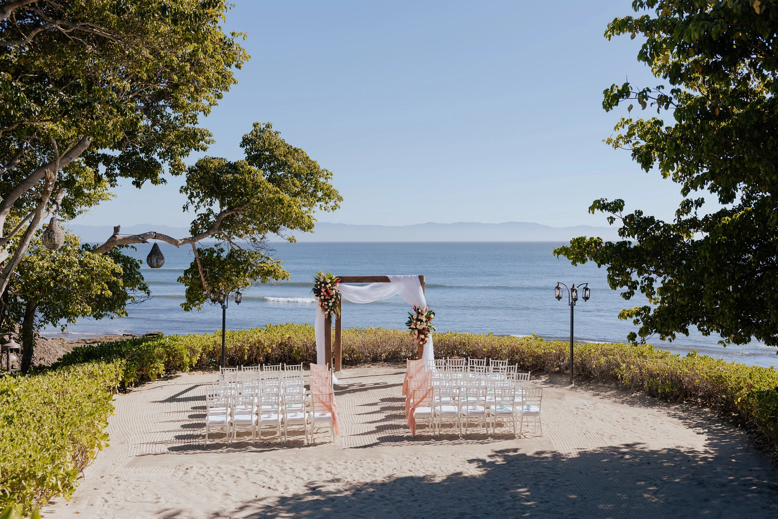 drbmisandterracewedd03682386df4bcb2 be all inclusive Beach wedding ceremony setup with a wooden arch draped in white fabric and pink florals overlooking the ocean. White chairs with pink sashes line a sandy aisle.
