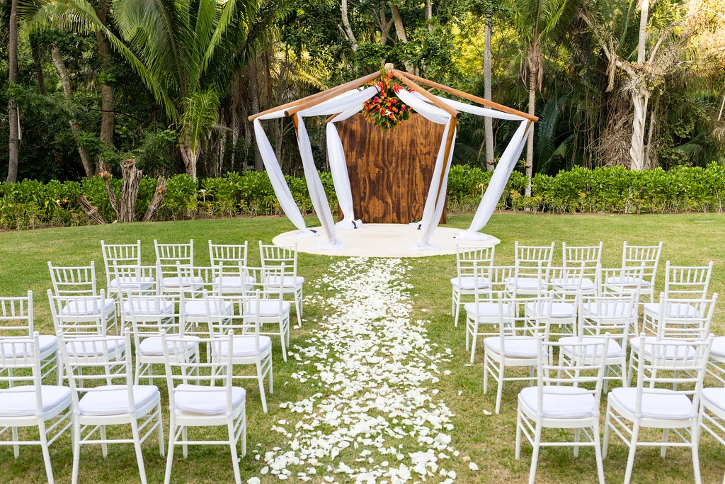 i2h2s8l3xljpg683f5af8d0de4 be all inclusive Outdoor wedding ceremony setup with white chairs along a petal-strewn aisle toward a wooden arbor draped in white fabric and flowers.