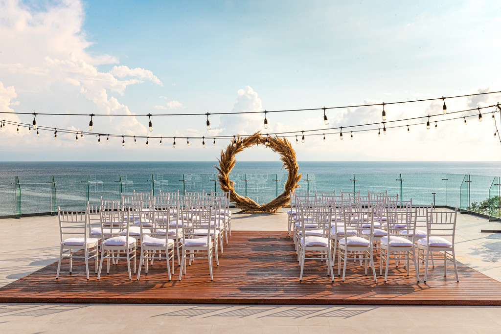 i2kbpfh2xljpg be all inclusive Outdoor seaside wedding ceremony setup with white chairs on a wooden aisle, a circular woven arch, string lights overhead, and an ocean view.