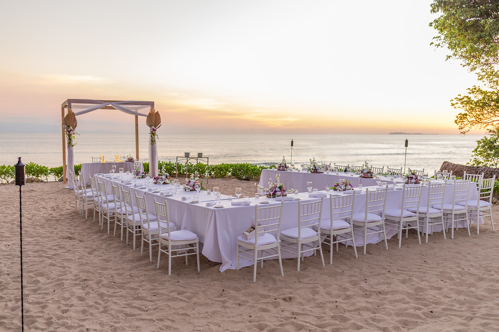 ib9gs5lgxljpg be all inclusive Beach wedding setup on white sand: long U-shaped tables, white chairs, floral centerpieces, under a wooden arch at sunset by the ocean.