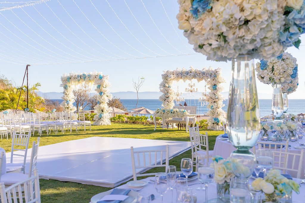 ibkjvgt3xljpg be all inclusive Outdoor seaside wedding venue with white chairs, floral arches, and tables set with white linens and blue accents overlooking the ocean.