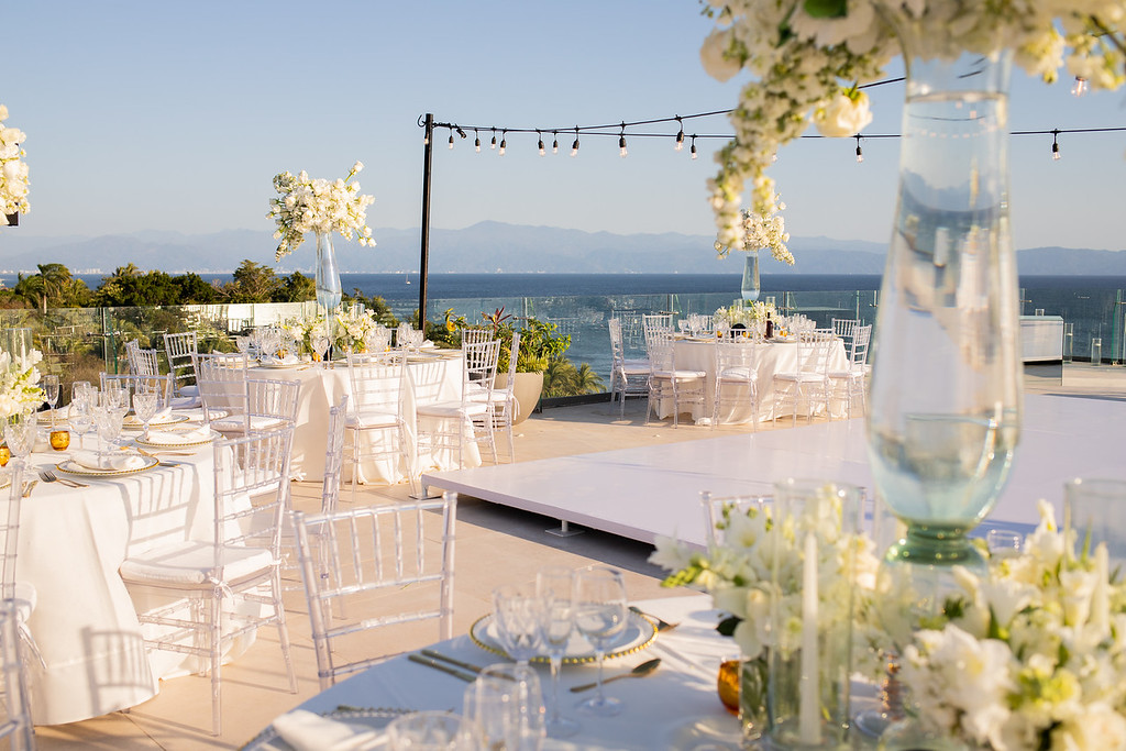 if4vxcvlxljpg be all inclusive Elegant seaside outdoor wedding reception with white tables, clear chairs, string lights, and an ocean view behind.