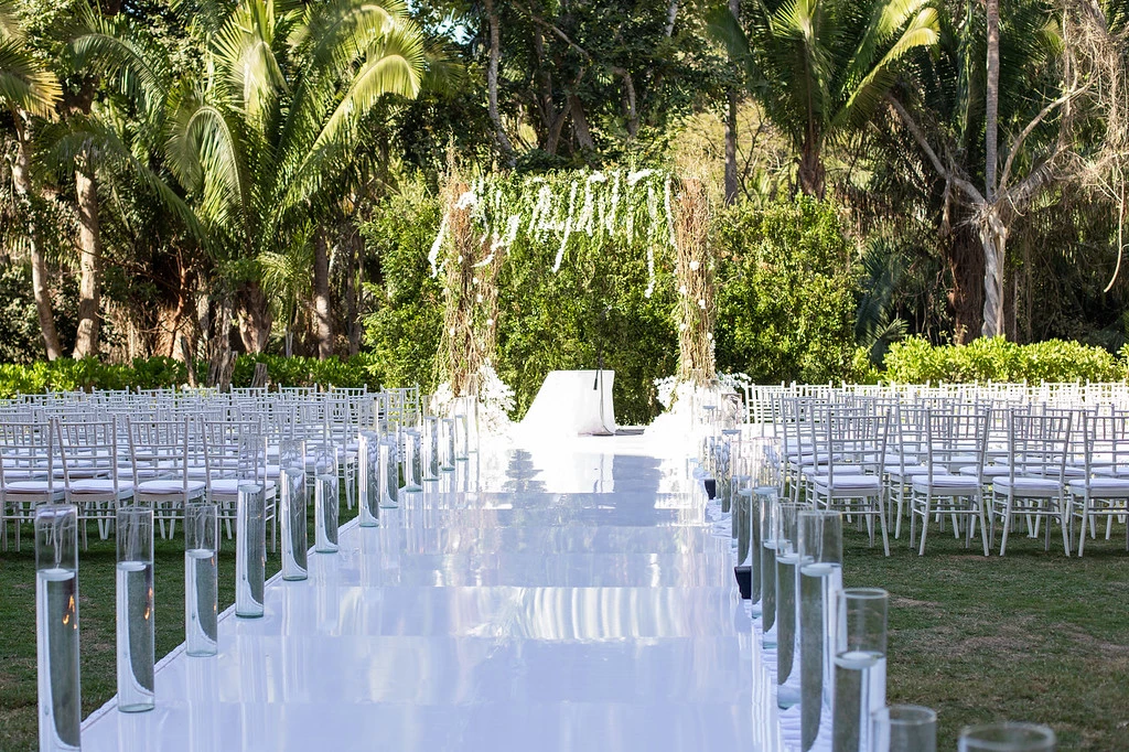 ib6vmk2sxljpg683f4b8a809a7 be all inclusive Outdoor wedding ceremony setup with a white aisle, glass candle holders along both sides, and a floral arch at the far end in a tropical garden.