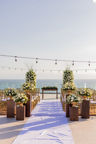 ibrbs4kxljpg be all inclusive Outdoor seaside wedding ceremony setup with white floral arrangements along a white aisle runner and wooden chairs facing the ocean.