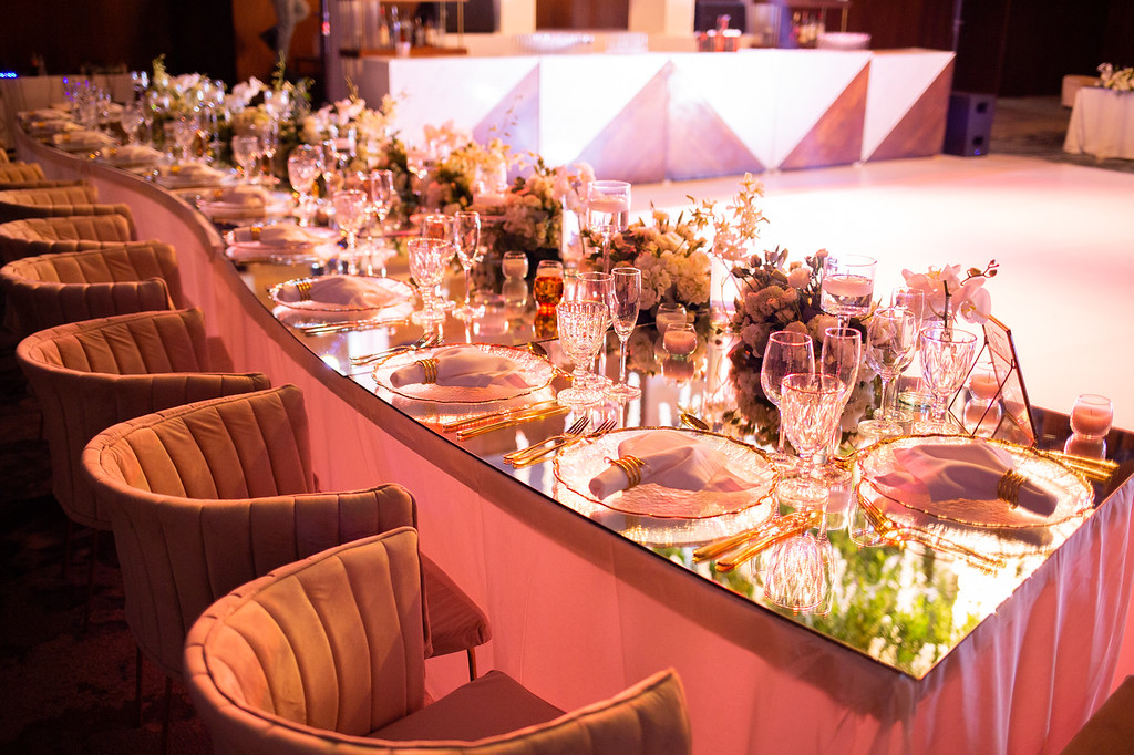 idvw5knkxljpg be all inclusive Elegant banquet table with mirrors, crystal glassware, white napkins, and floral centerpieces; curved row of upholstered chairs with pink lighting in the background.