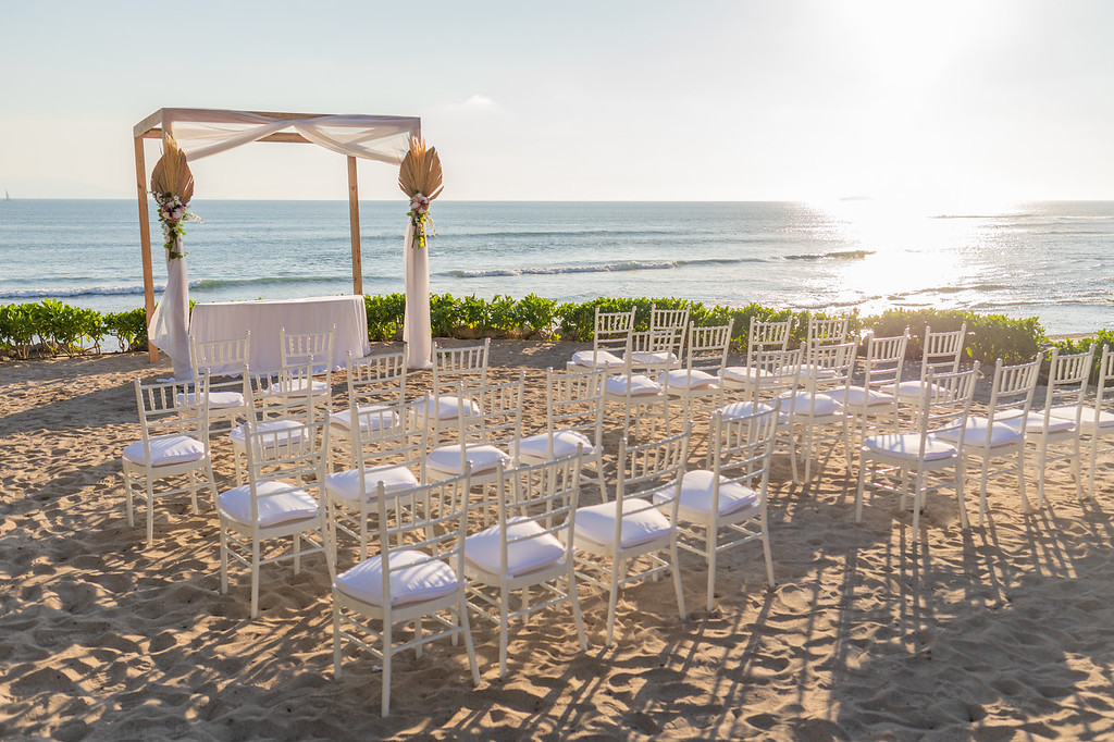 ih2vp853xljpg be all inclusive Beach wedding ceremony setup with white chairs facing a wooden arch draped in white fabric, overlooking the ocean.