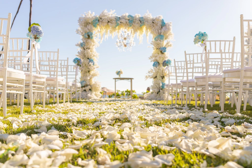ih5sqzbzxljpg683f5af0844c4 be all inclusive Outdoor wedding aisle with white chairs and a floral arch, petals scattered on the grass path