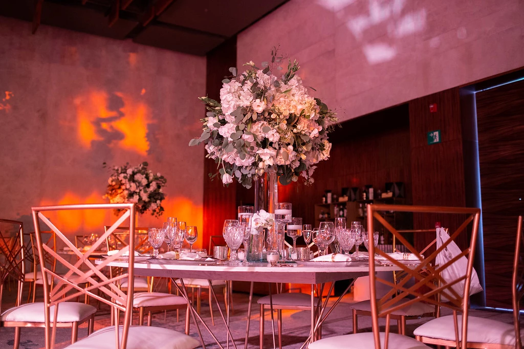 ihx9wdbvxljpg683f4b81d7212 be all inclusive Round banquet table with a tall white floral centerpiece, crystal glassware, and rose-gold chairs in a warmly lit event space.