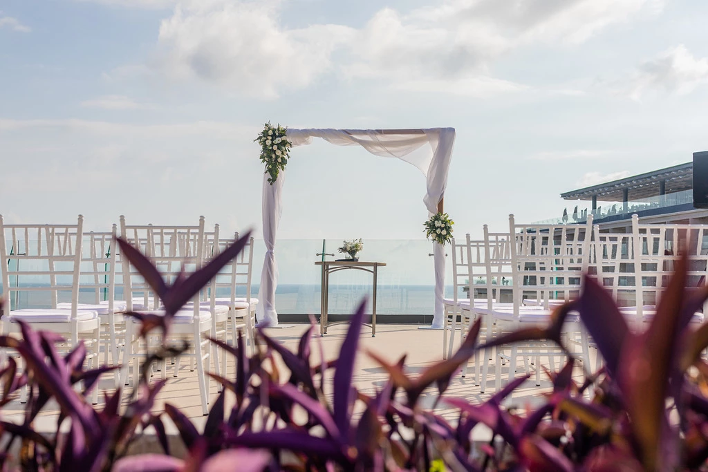 im7gq9bgxljpg683f5b39c6c48 be all inclusive Outdoor seaside wedding setup on a rooftop terrace with a white-draped arch, floral arrangements, and rows of white chairs facing a small altar table; ocean in the background.