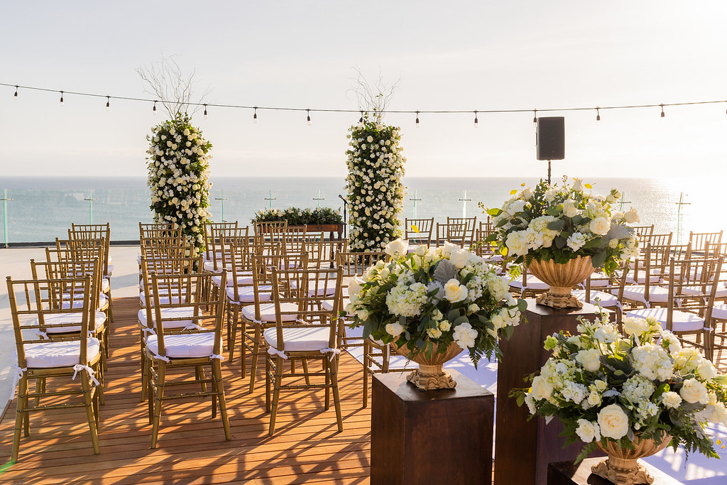 ipbktkfqxljpg be all inclusive Outdoor wedding ceremony setup on a wooden deck by the sea with gold chairs and white floral arrangements under string lights.