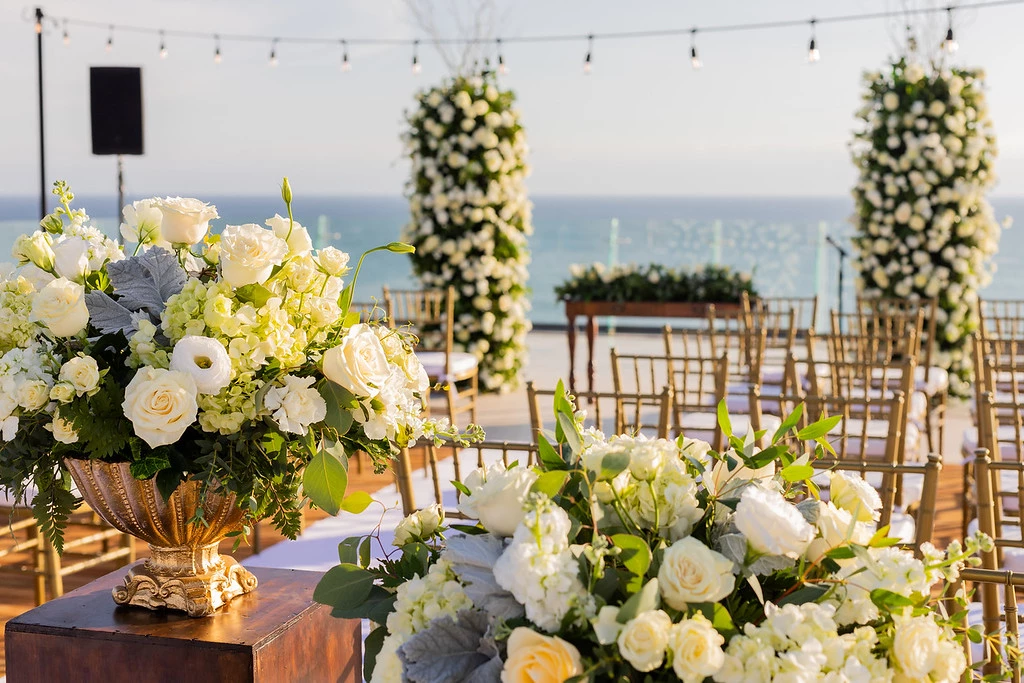 ipfprxc7xljpg683f5af6b9158 be all inclusive Elegant seaside wedding ceremony setup with white and cream flowers in ornate urns, gold chairs, and string lights overlooking the ocean.