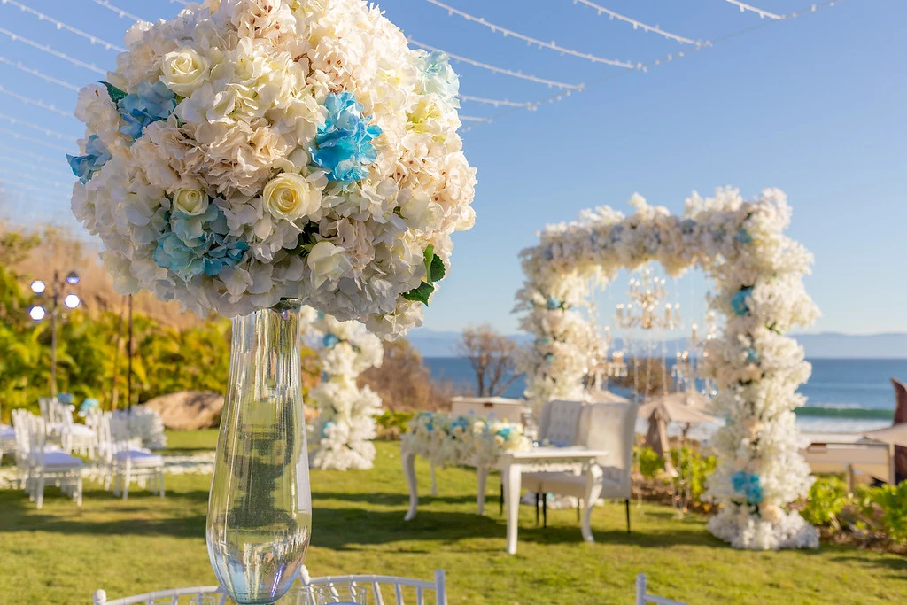 ipvmmqzdxljpg683f5af2bc626 be all inclusive Tall glass vase holds a large white hydrangea bouquet with blue accents at an outdoor beach wedding setup.