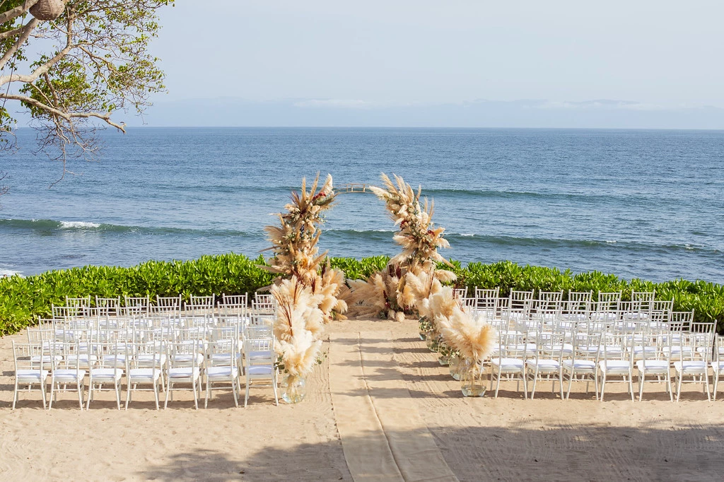 isgzgxbrxljpg683f4b7acc5fa be all inclusive Beach wedding ceremony setup with white chairs facing a flower arch overlooking the ocean.