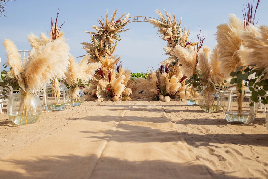 ivz9hd5bxljpg683f4b79a955e be all inclusive Outdoor beach wedding setup with pampas grass arches and a sandy aisle leading to a floral arch ceremony backdrop.