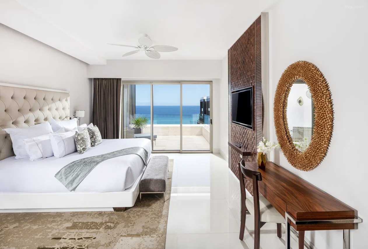 0220p12000k7e3mhg3107w1280853r5 be all inclusive Hotel bedroom with a king bed, tufted headboard, and a view of the ocean through sliding glass doors to a balcony.