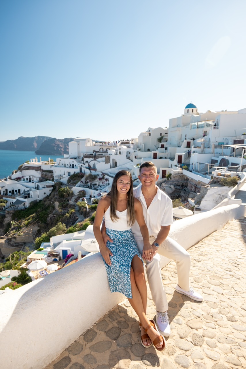 Couple sitting on a white wall in a sunny Santorini town with blue domed church in background, smiling at the camera.