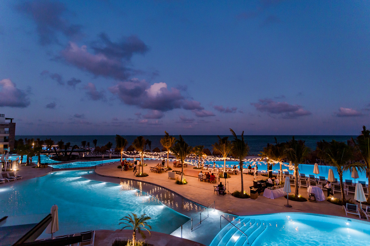 1 be all inclusive Resort pool area at dusk with illuminated pools, palm trees, and a seaside view.