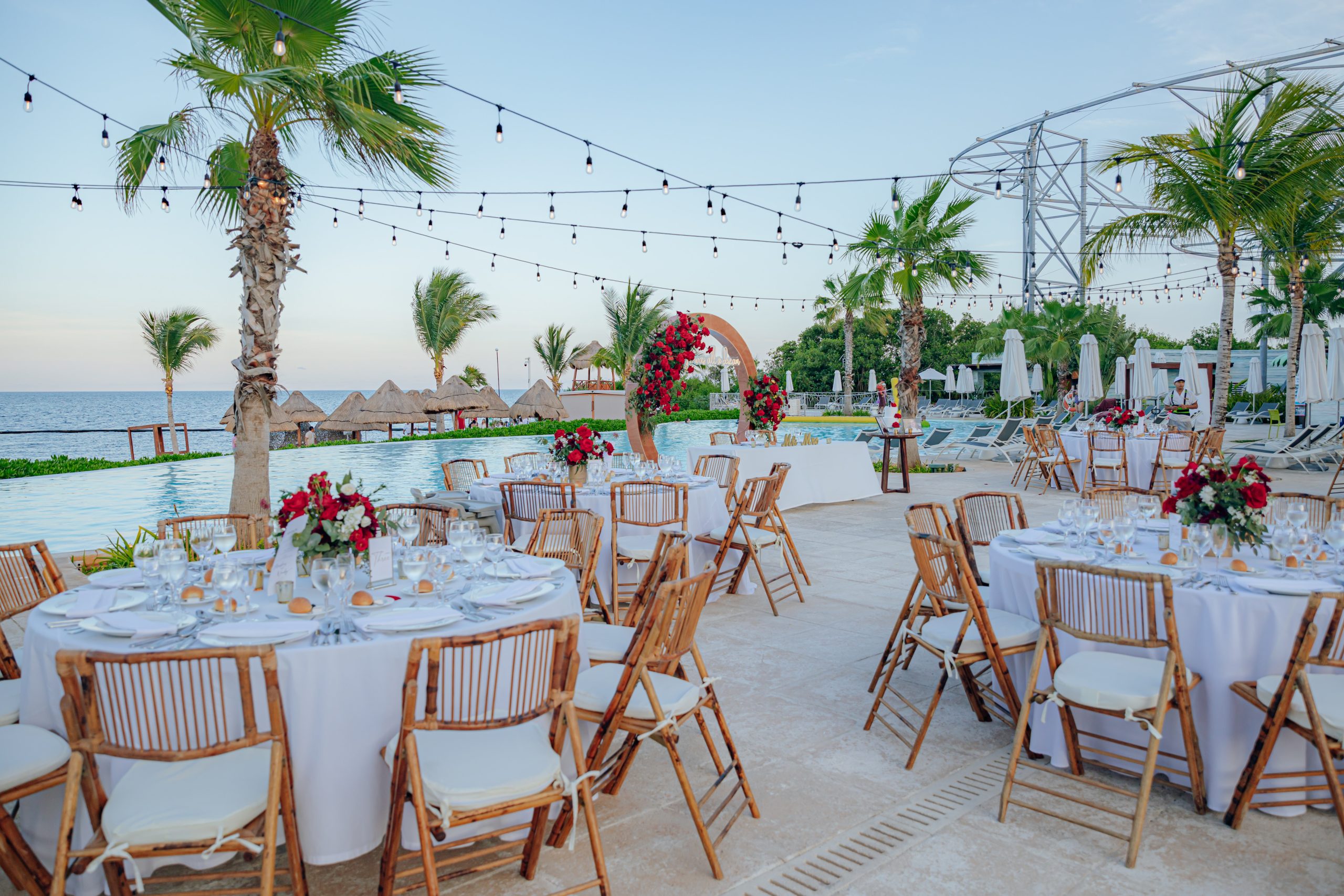 markus wendy vickers be all inclusive Oceanfront wedding reception with round tables, white cloths, and red floral arch by the pool under string lights.