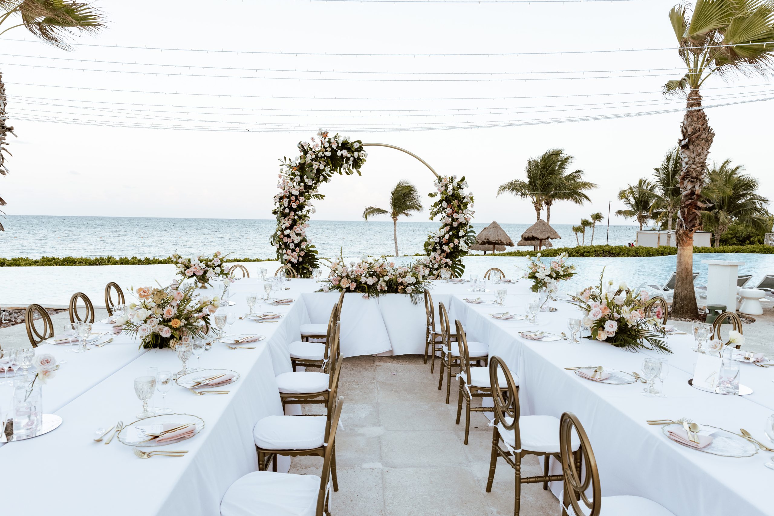 jaqueline michael be all inclusive Outdoor beach wedding setup with long white-draped tables, gold chairs, and a floral arch by the sea