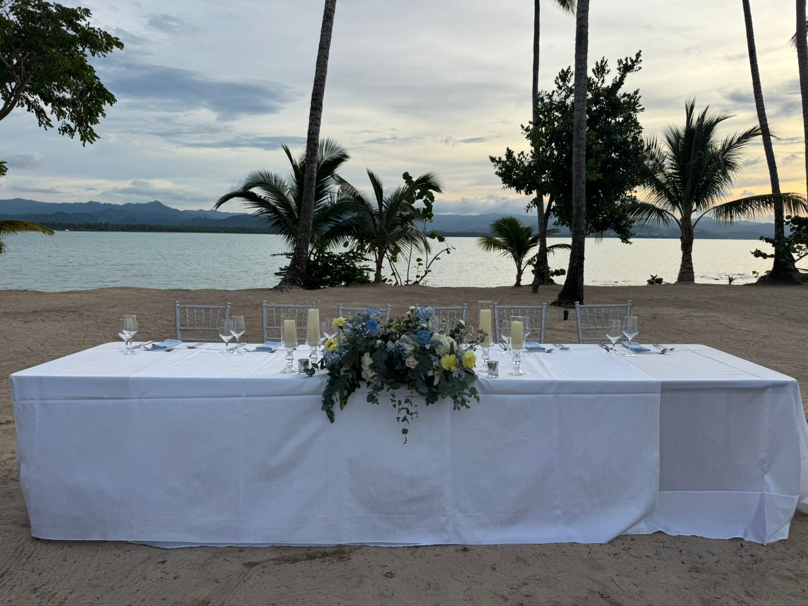 1bb57db4ac904f7d8ea04598f8e431e1 be all inclusive Beach wedding table setup on sand with white linen, floral centerpiece, candles, and glassware against calm sea and palm trees at sunset.