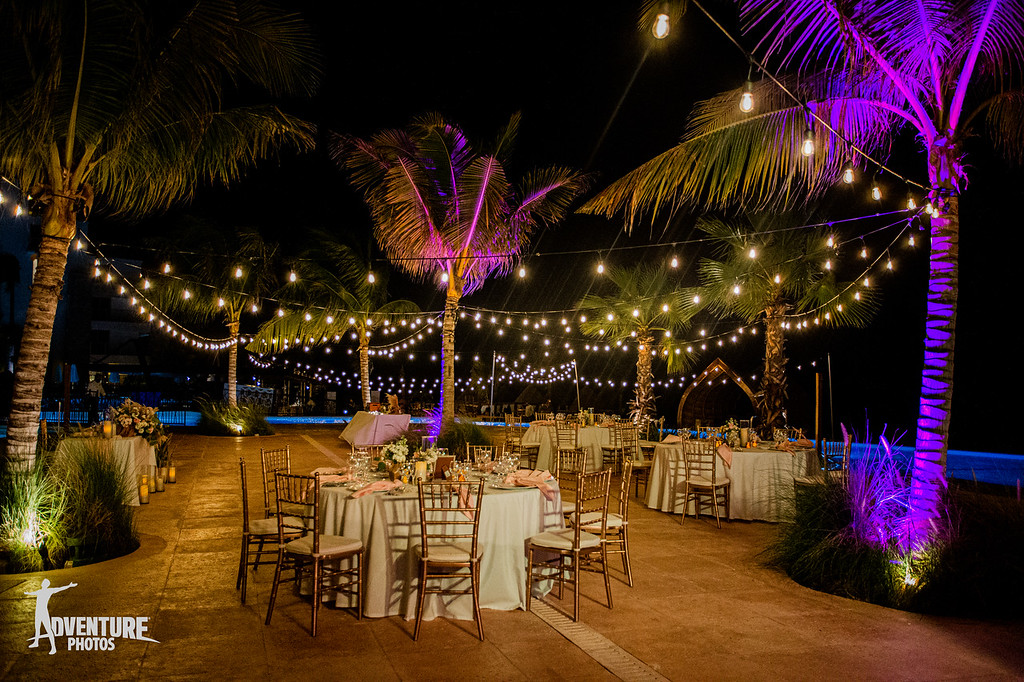 2 be all inclusive Night outdoor event setup: round tables with gold chairs under string lights between palm trees by a pool.