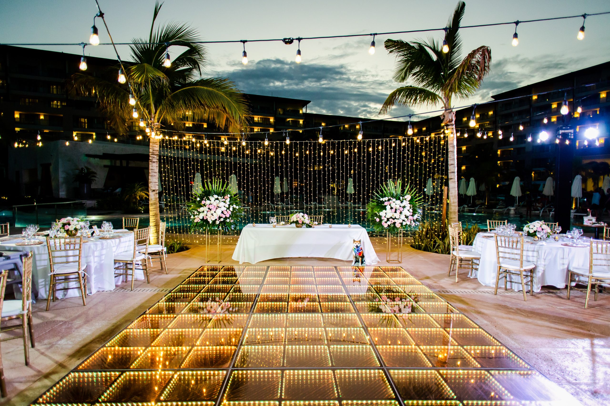 6 be all inclusive Outdoor evening wedding reception with string lights, palm trees, and a glowing glass-tile dance floor by a pool.