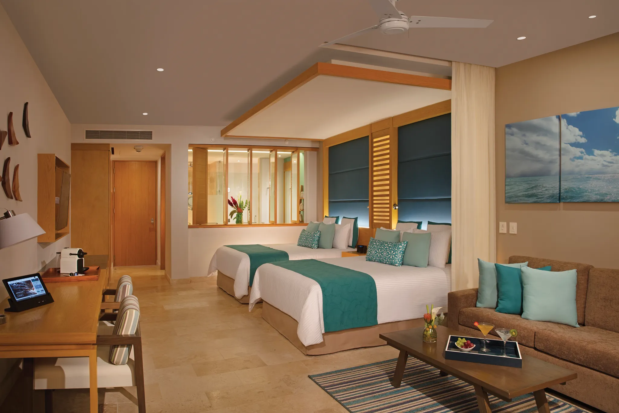 drepmjrdouble1acb be all inclusive Hotel suite with two queen beds, teal bed runners and pillows, and a seating area with a sofa and coffee table.