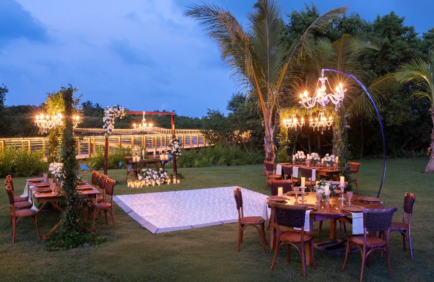 screenshot 20260707 093822 be all inclusive Outdoor evening wedding reception with a glowing square dance floor, chandeliers, and round tables under string lights on a lawn.