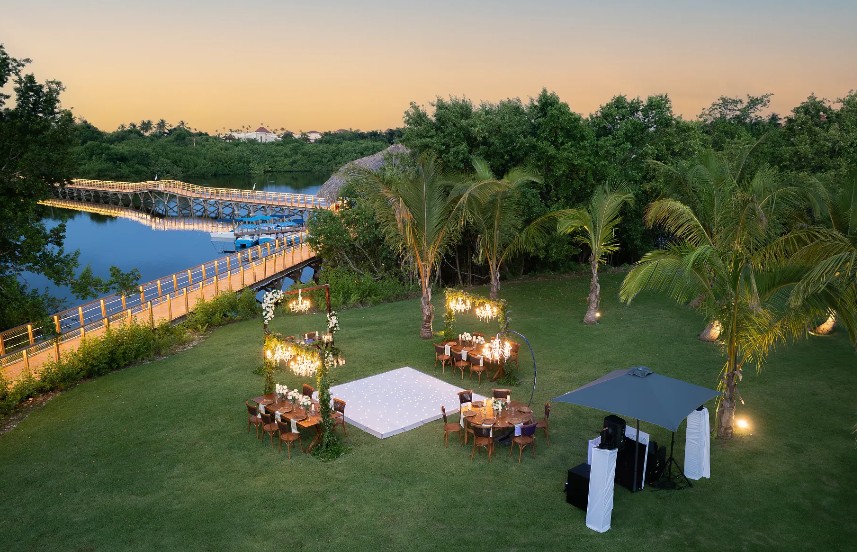 screenshot 20260707 093859 be all inclusive Outdoor wedding reception at dusk on a grassy lawn beside a river; illuminated square dance floor, round tables, and palm trees nearby.