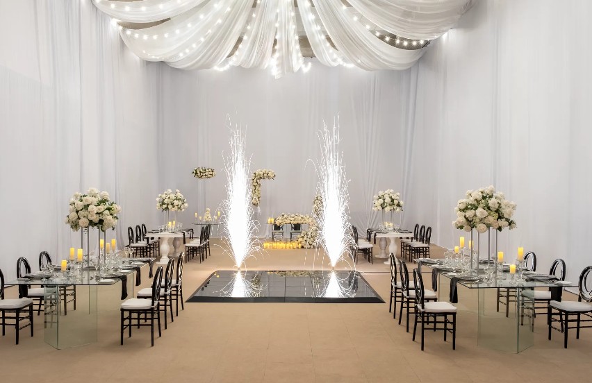 screenshot 20260707 093919 be all inclusive Elegant wedding reception hall with white drapes, floral centerpieces, and a glowing fountain centerpiece on a mirrored platform across two long glass-topped tables.