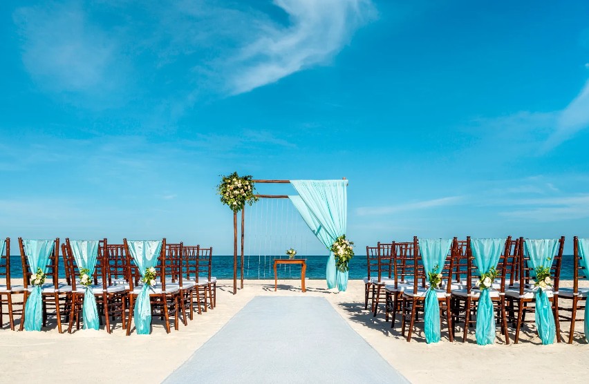 screenshot 20260707 093934 be all inclusive Beach wedding setup with a teal-draped arch and rows of chairs facing the ocean. A light aisle runs toward the altar.