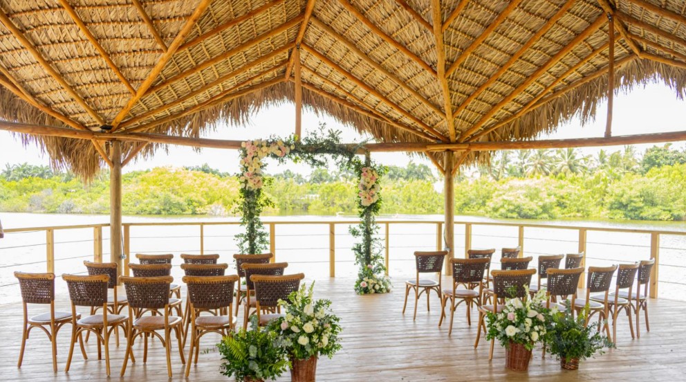 screenshot 20260707 094229 be all inclusive Outdoor wedding ceremony setup on a wooden deck beneath a thatched roof, with a floral arch and rows of wicker chairs facing the water.