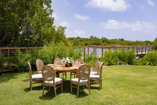 screenshot 20260707 094250 be all inclusive Outdoor lawn dining setup with a round wooden table and six wicker chairs, floral centerpiece, by a lake with a wooden railing nearby.