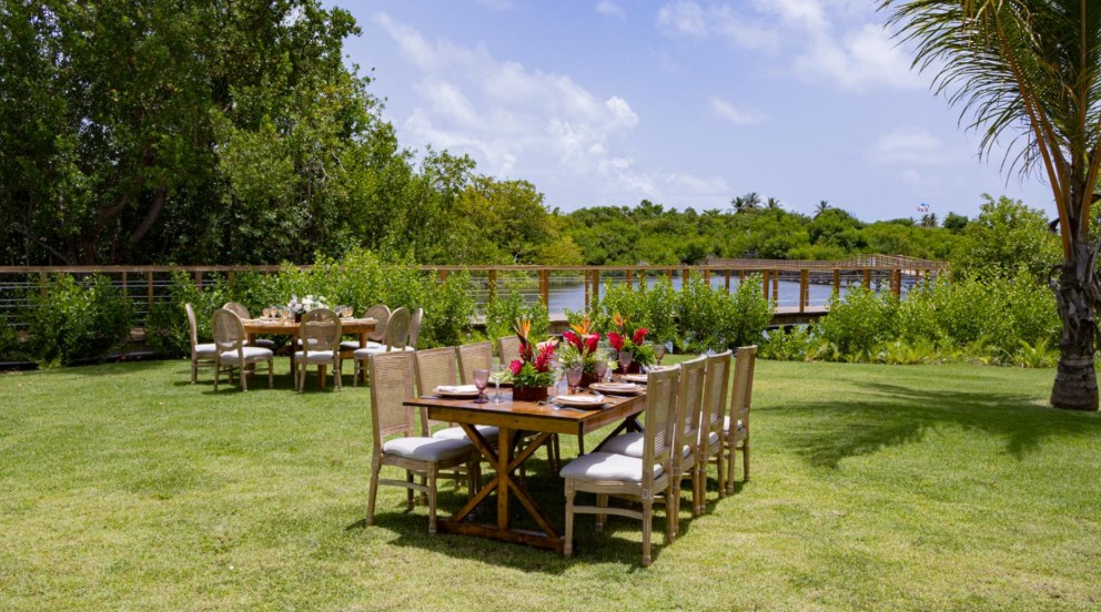 screenshot 20260707 094259 be all inclusive Outdoor dining setup with two wooden tables and chairs on a grassy lawn by a lake, surrounded by lush greenery and a wooden boardwalk in the background.