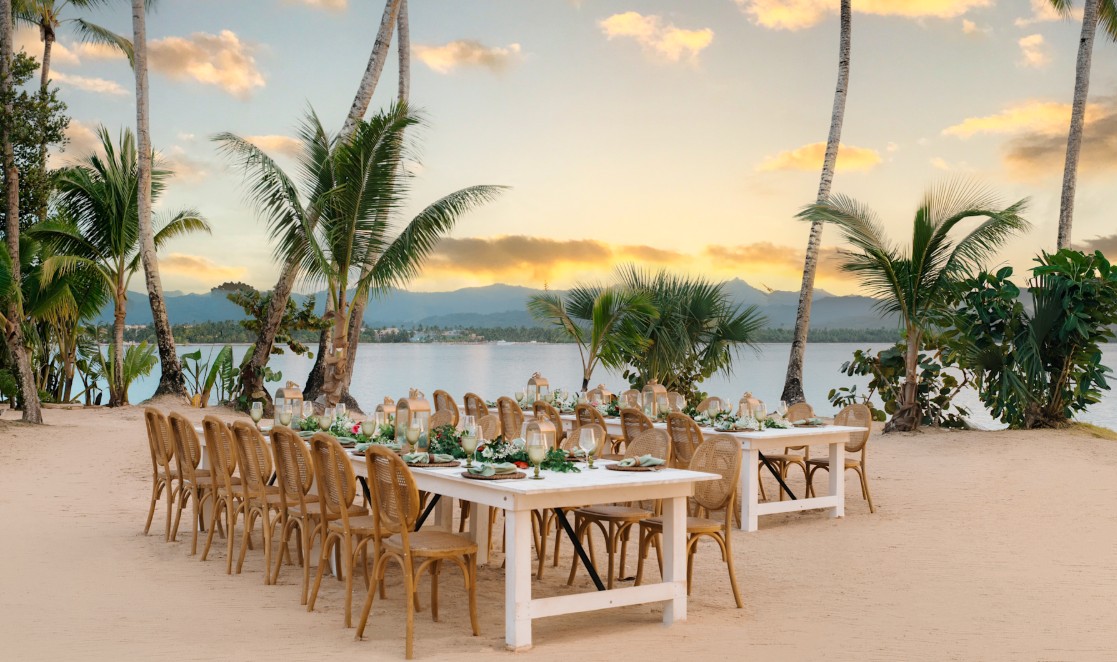 screenshot 20260710 131154 be all inclusive Long white dining tables set for a banquet on a sandy beach, surrounded by wicker chairs, lanterns, greenery, and palm trees at sunset.