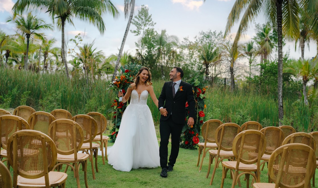 screenshot 20260710 131209 be all inclusive Bride in a strapless white gown and groom in a black suit walk hand in hand down an outdoor aisle lined with wicker chairs and a tropical flower arch behind them, with palm trees and greenery in the background.
