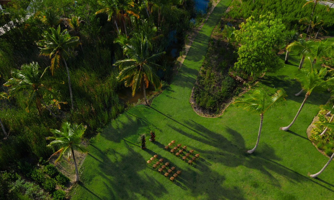 screenshot 20260710 131235 be all inclusive Aerial view of a tropical garden with rows of wooden chairs arranged for an outdoor ceremony.