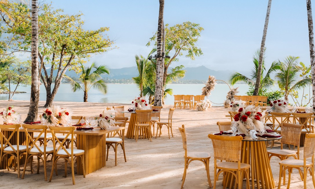 screenshot 20260710 131248 be all inclusive Outdoor beachside dining setup with wooden tables and chairs, tropical trees, and floral centerpieces by calm water.