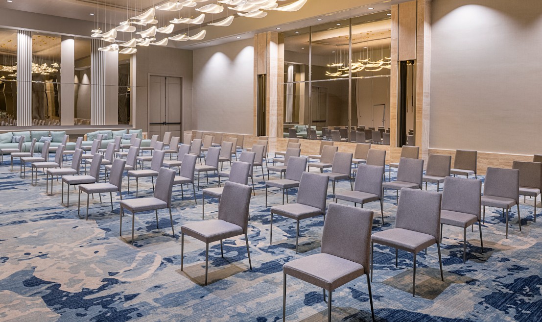 screenshot 20260710 131308 be all inclusive Rows of gray chairs set up for a conference in a spacious hotel ballroom with blue patterned carpet and modern lighting.