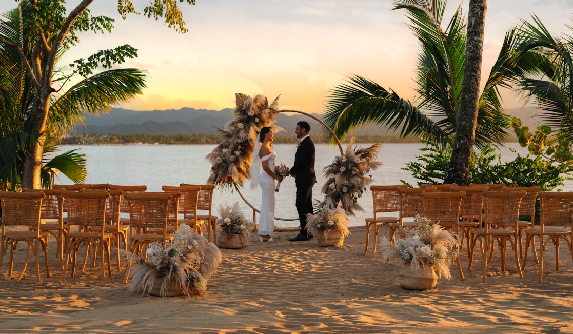 screenshot 20260710 131318 be all inclusive Bride and groom exchange vows under a circular floral arch on a sandy beach at sunset, with rows of wooden chairs and tropical palms nearby.
