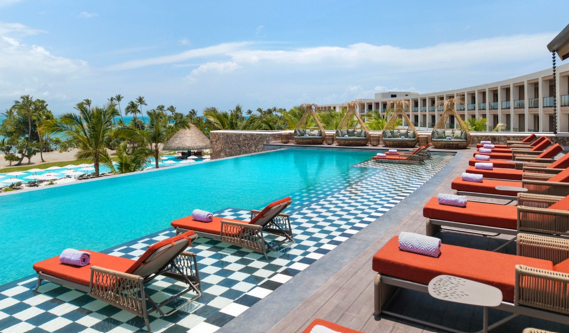 screenshot 20260710 133141 be all inclusive Luxurious resort poolside scene with orange lounge chairs, rolled towels, and checkered pool deck facing a turquoise pool and palm-lined beach.