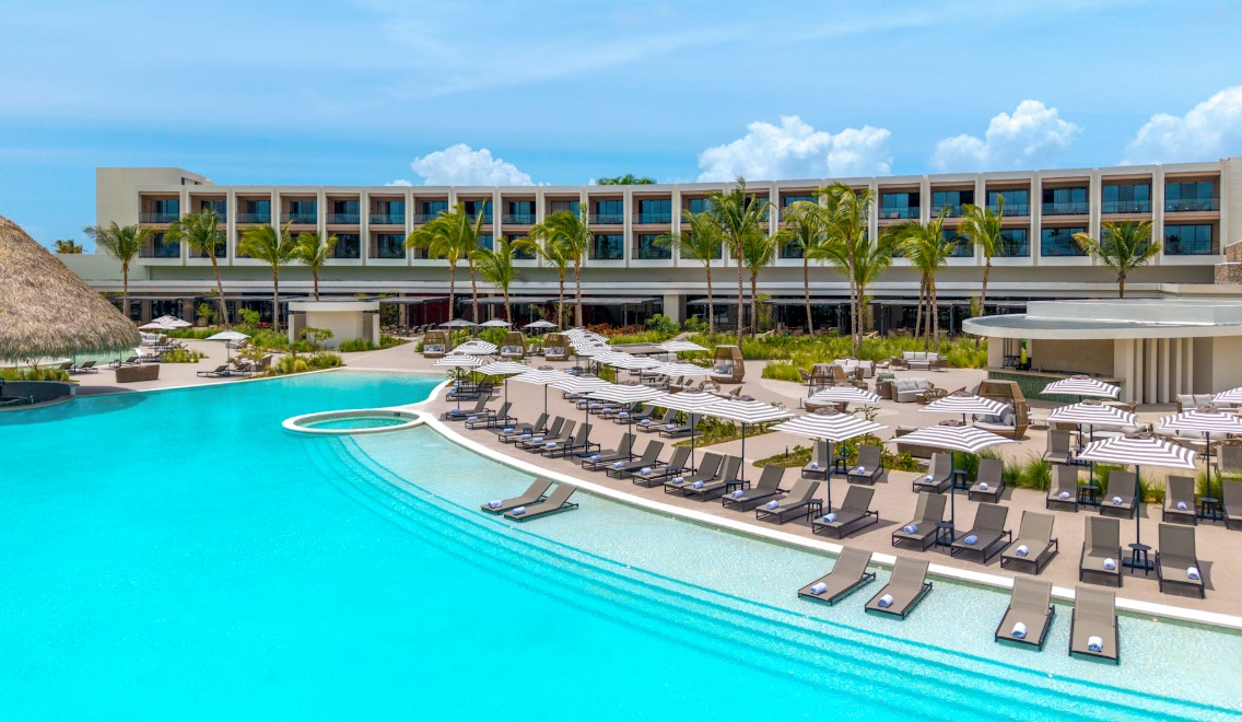screenshot 20260710 133155 be all inclusive Resort pool area with rows of lounge chairs and striped umbrellas, palm trees, and a modern hotel building in the background.