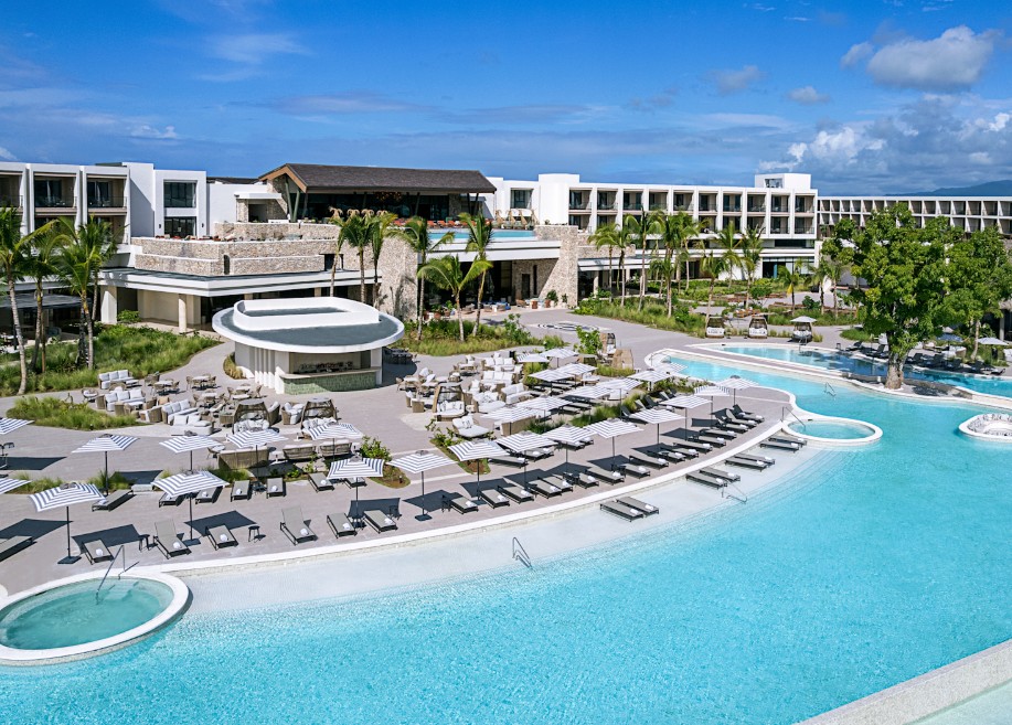 screenshot 20260710 133435 be all inclusive A luxury resort pool area with curved pool, loungers under striped umbrellas, and palm trees, set against white hotel buildings.