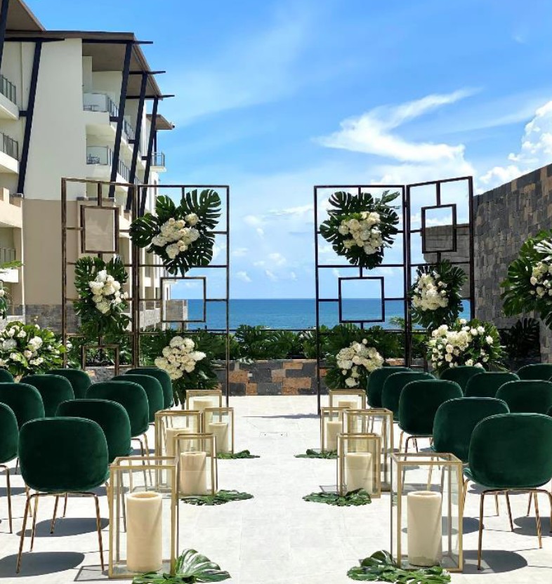 screenshot 20260713 155346 be all inclusive Outdoor seaside wedding setup with green chairs, white flowers, and lanterns along a white aisle overlooking the ocean.