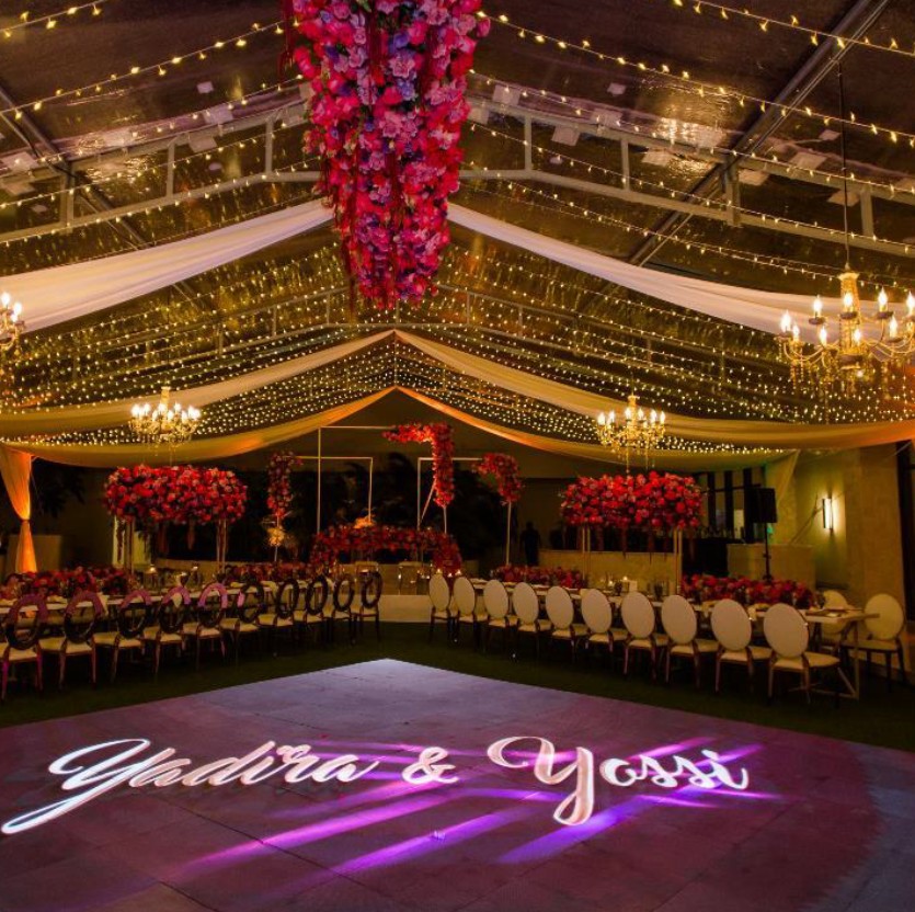 screenshot 20260713 155539 be all inclusive Wedding reception inside a decorated tent with hanging lights, chandeliers, and tall floral centerpieces, floor shows glowing names 'YudhiA & Yoss' projected.