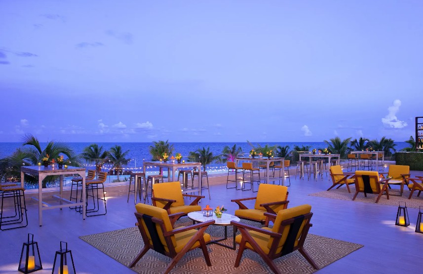 screenshot 20260713 164015 be all inclusive Outdoor beachfront lounge with yellow-cushioned chairs and wooden tables on a terrace, string lights, and palm trees against the ocean at dusk.