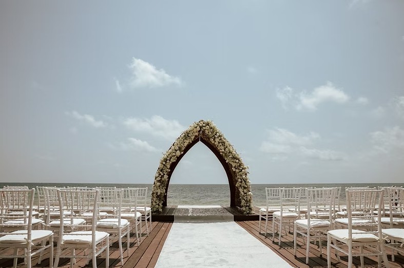 screenshot 20260713 164212 be all inclusive Flower-covered wedding arch at a beach ceremony, white chairs arranged along a wooden aisle facing the arch by the sea.